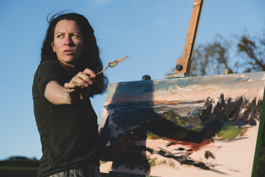 Women artist holding a paint brush stood in front of a picture on an easel