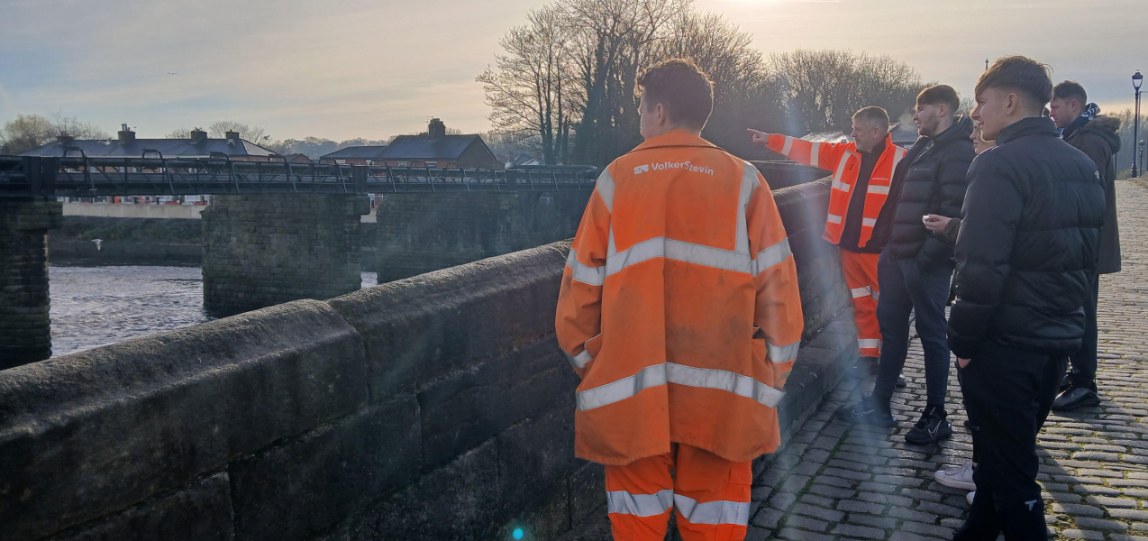 Group  of men and women stood on a bridge looking over the wall along the river